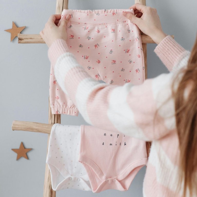 Person holding pink patterned shorts and a white bodysuit on a wooden ladder.