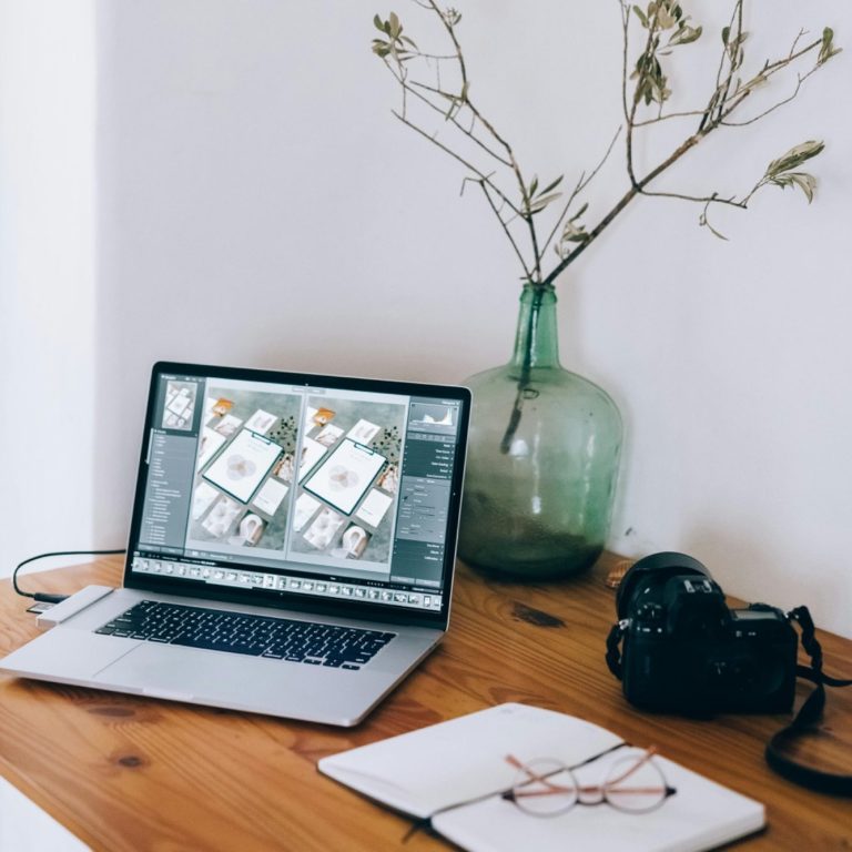 Laptop on a wooden desk with a camera, notebook, glasses, and a vase with dried branches.