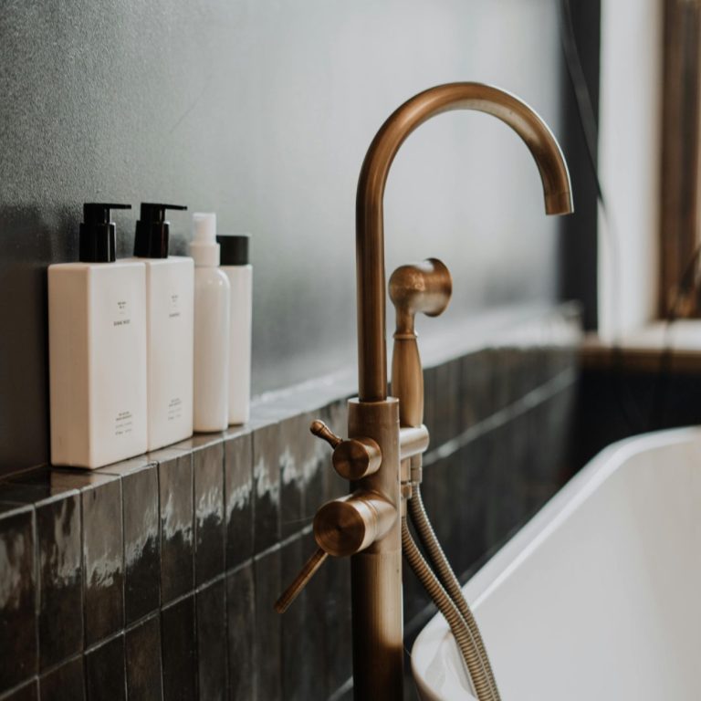 Bathroom makeover package Brass tap and white shampoo bottles beside a modern bathtub against a dark wall.