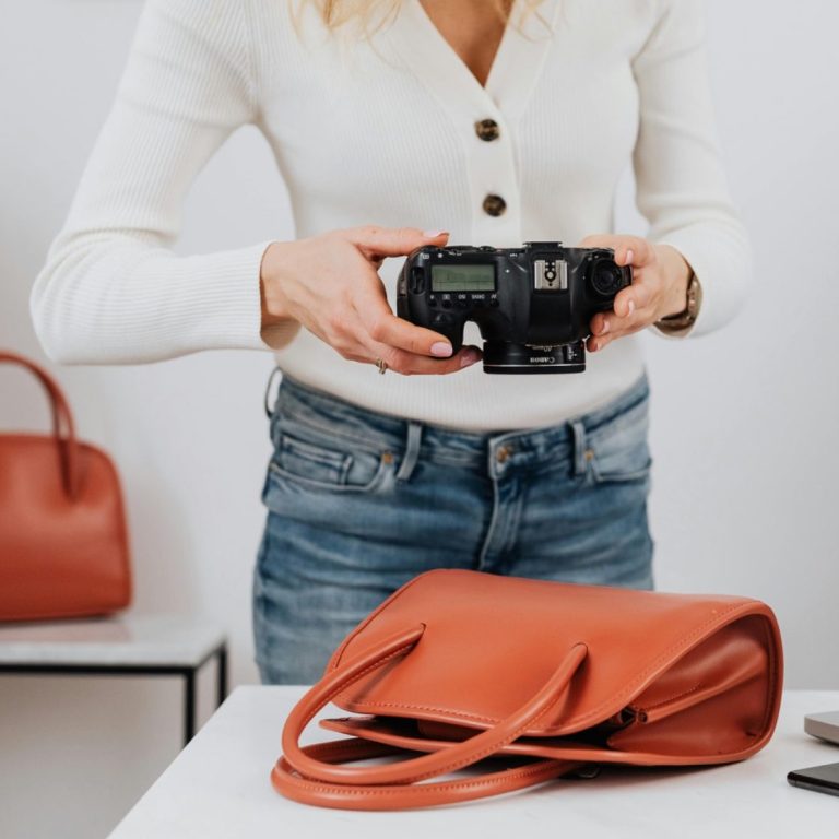Professional product photography services Person holding a camera beside a brown handbag on a table.