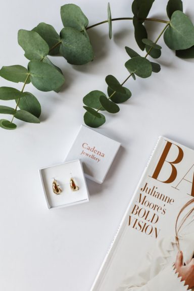 Gold earrings presented in a white box, accompanied by a magazine and greenery.