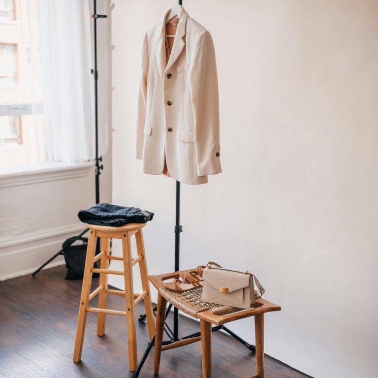 Beige blazer hanging on a rack beside a wooden stool and a small handbag.