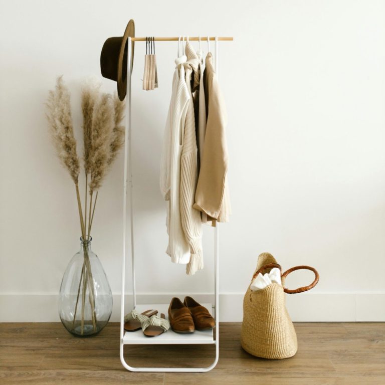 A minimalist clothing rack with hats, garments, and a woven bag beside dried flowers.
